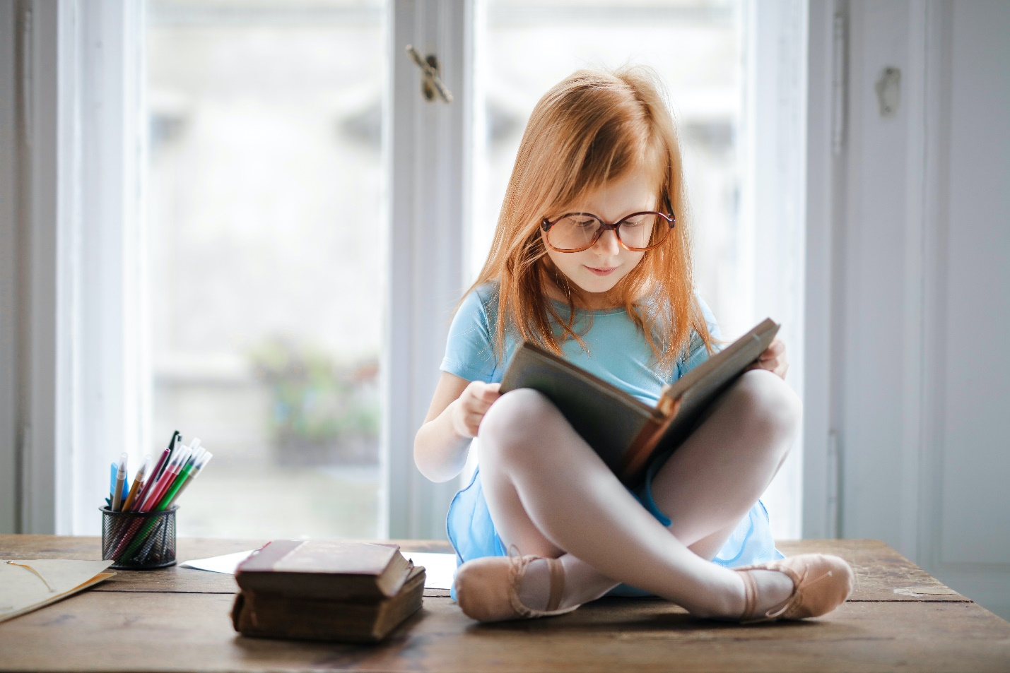 A young girl child sits cross-legged reading on a table beside two stacked books and a holder full of markers
