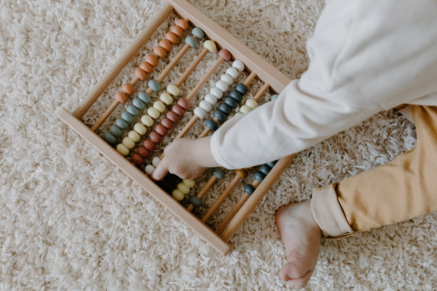 A toddler's hand and foot are seen with a wooden abacus on a plush carpet.
